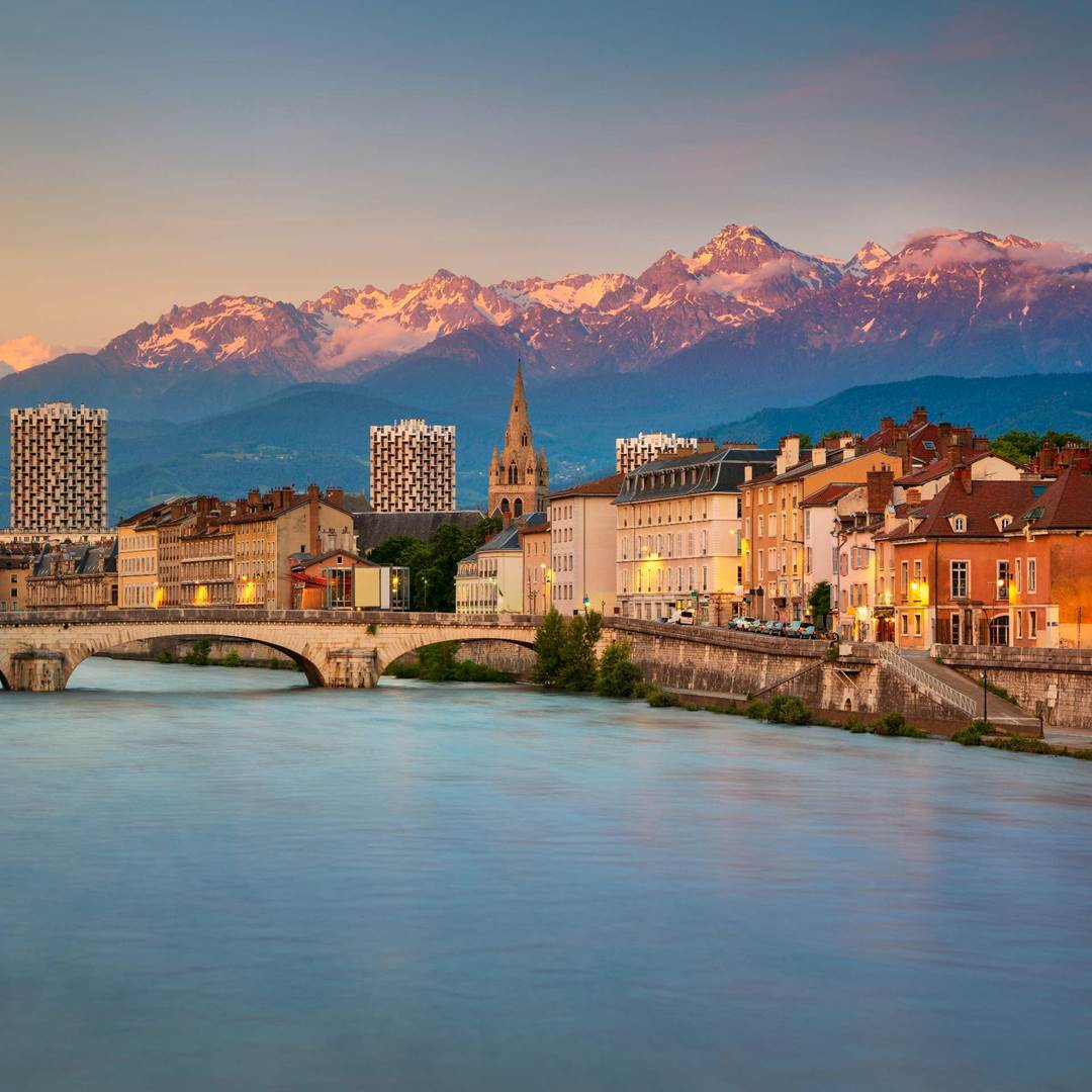 A river flows past colorful buildings and a stone bridge, with a church and snowy mountains in the background under a clear sky.