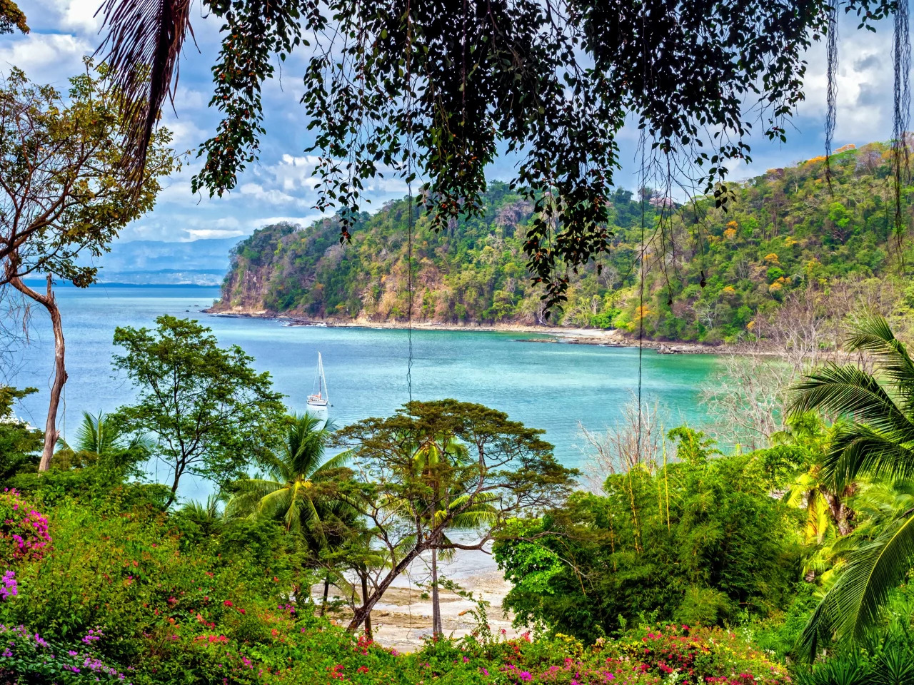 Tropical coastline with lush green trees, clear blue water, a sandy beach, and a sailboat anchored near the shore under a partly cloudy sky.
