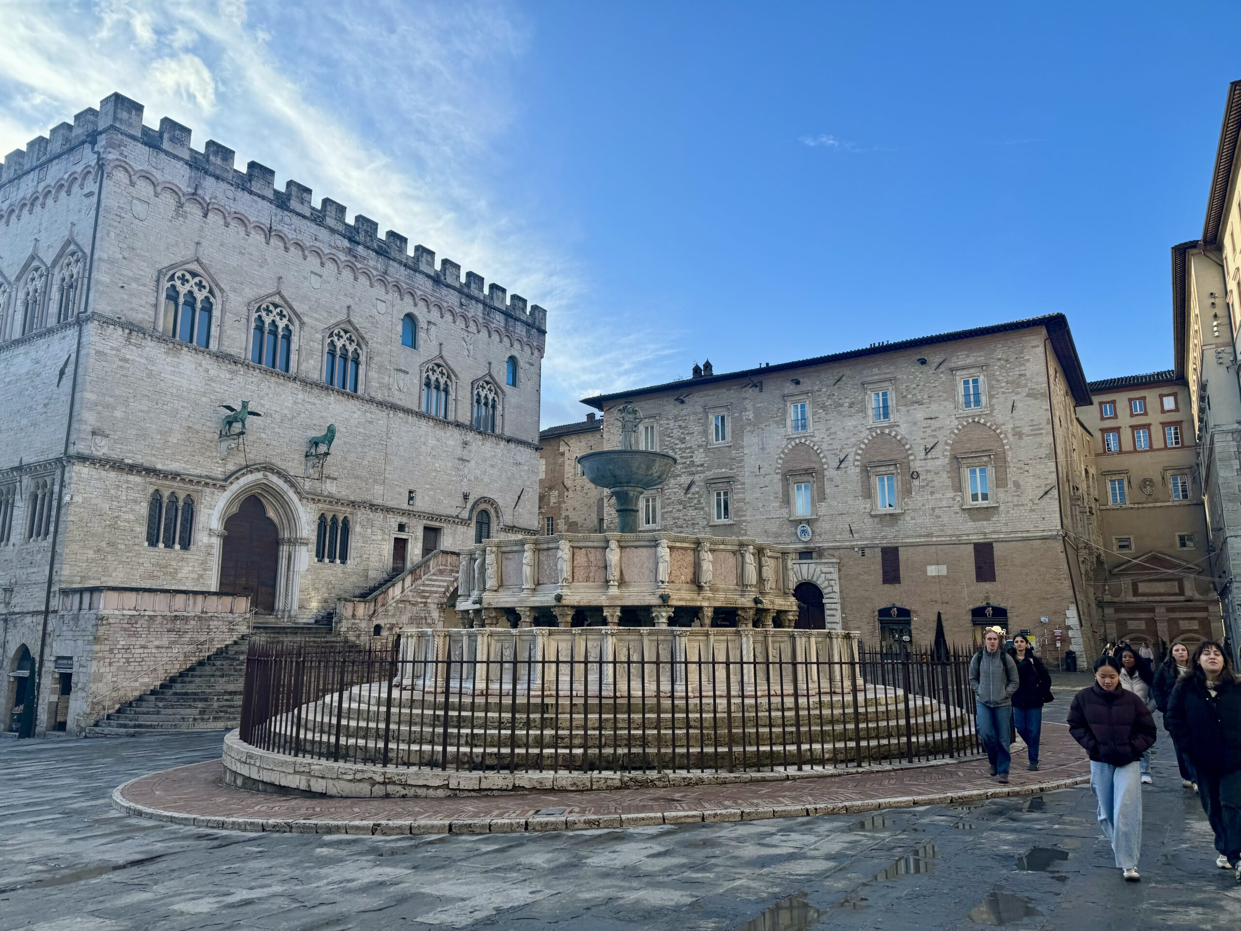 A stone fountain sits in a plaza surrounded by historic buildings, with a few people walking nearby under a partly cloudy blue sky.