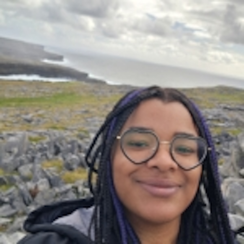 Person with glasses and dark hair smiles at the camera with rocky terrain, green grass, and a cloudy sky in the background.