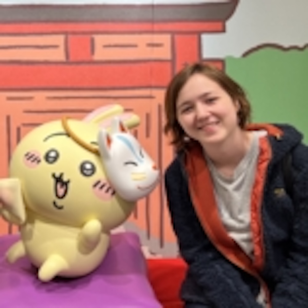 A person sits beside a large yellow cartoon character holding a white mask, against a colorful indoor backdrop.
