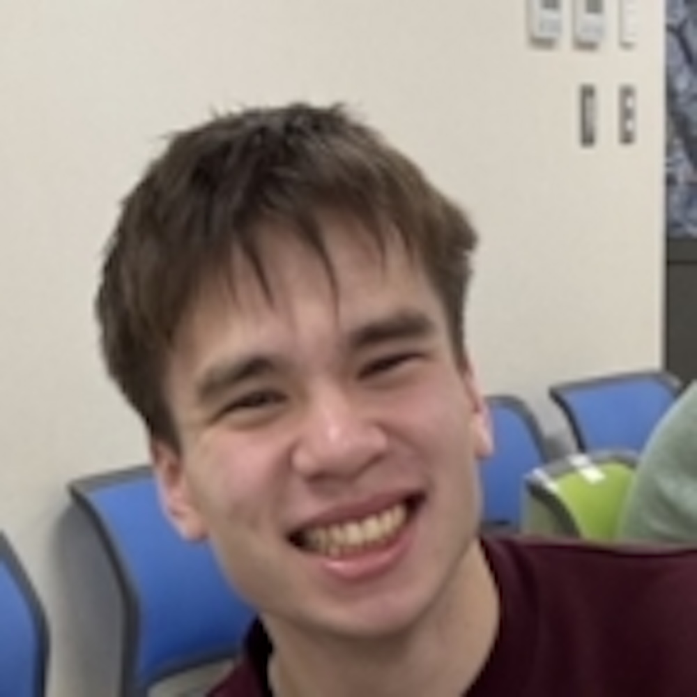 A young person with short brown hair smiles while seated indoors in a room with blue and green chairs.