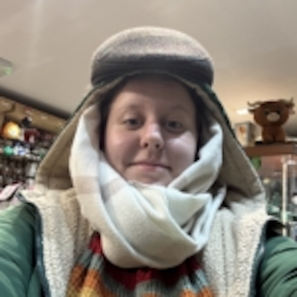 Person wearing a thick scarf, warm hat, and layered clothing indoors, with shelves and a plush deer head in the blurry background.