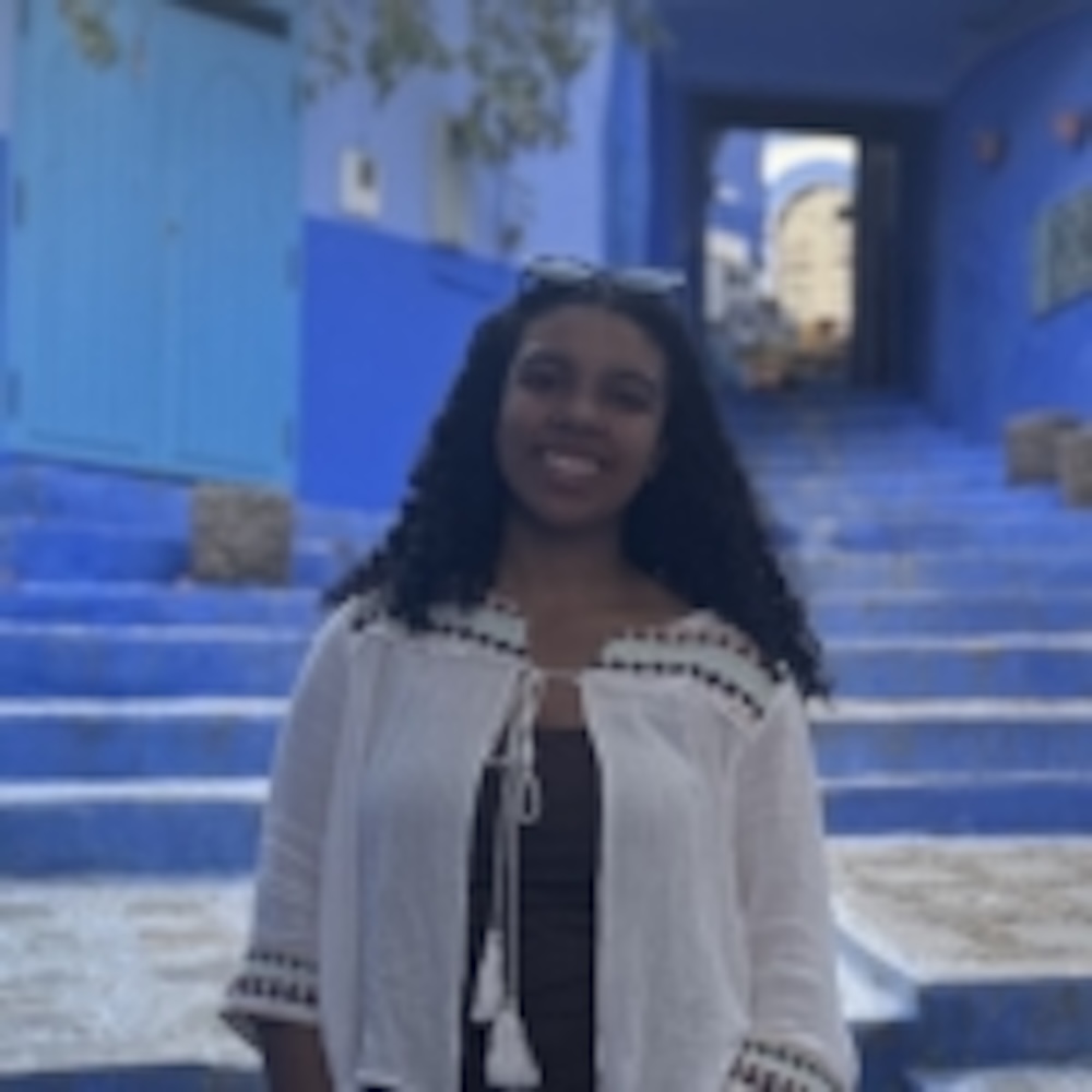 A woman with long curly hair stands on blue-painted steps in an outdoor area, wearing a white top and sunglasses on her head. Blue doors and walls are visible in the background.