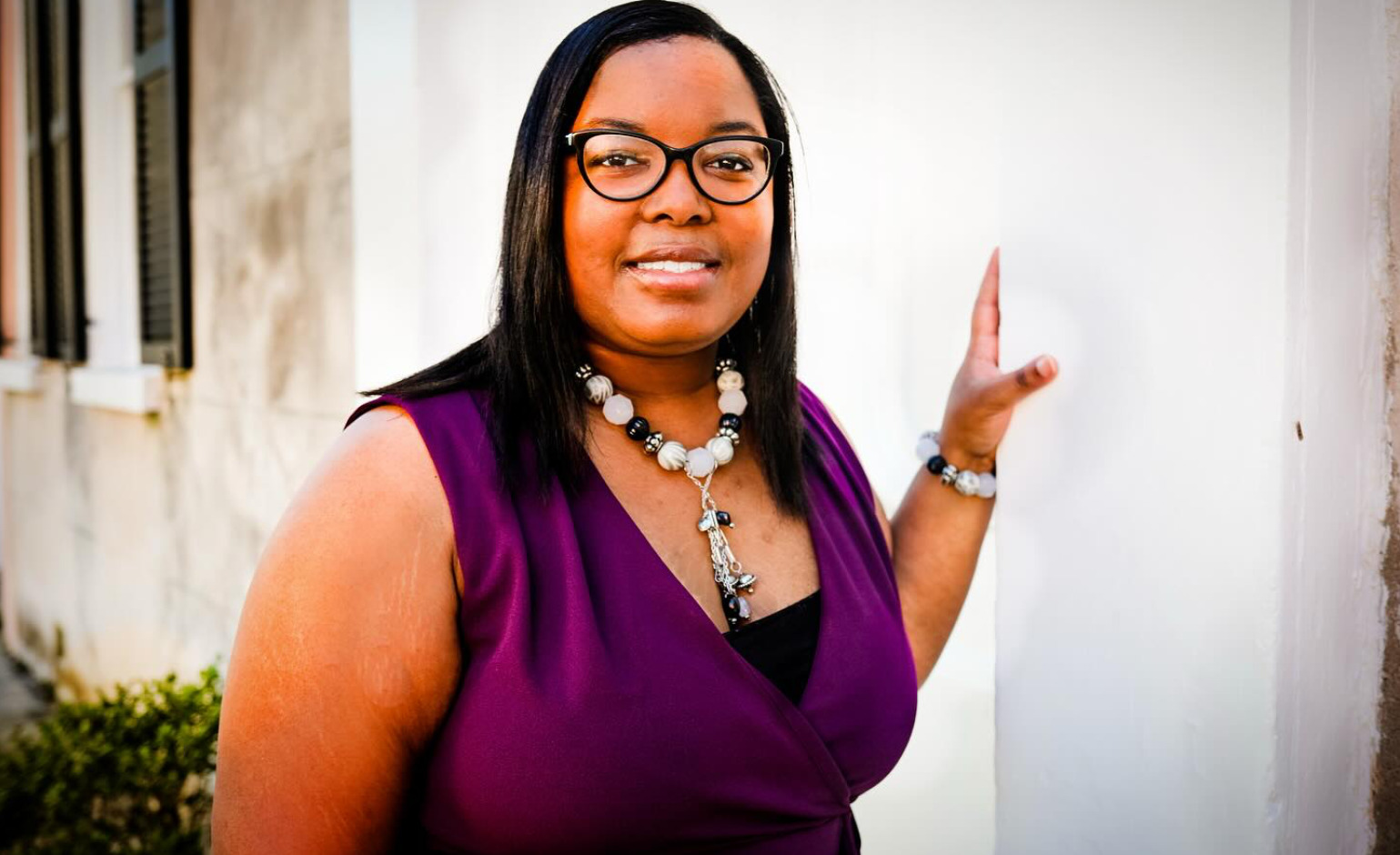 A woman with long dark hair and glasses, wearing a purple sleeveless top and a beaded necklace, stands with one hand touching a white pillar and smiles at the camera.