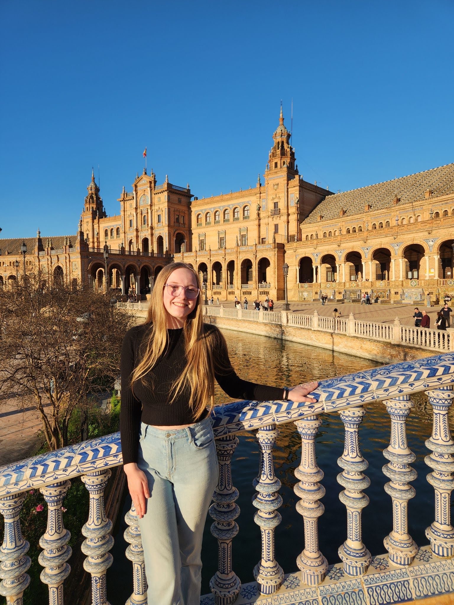 A person stands on a decorated bridge at Plaza de España in Seville, Spain, with an ornate building and clear blue sky in the background.