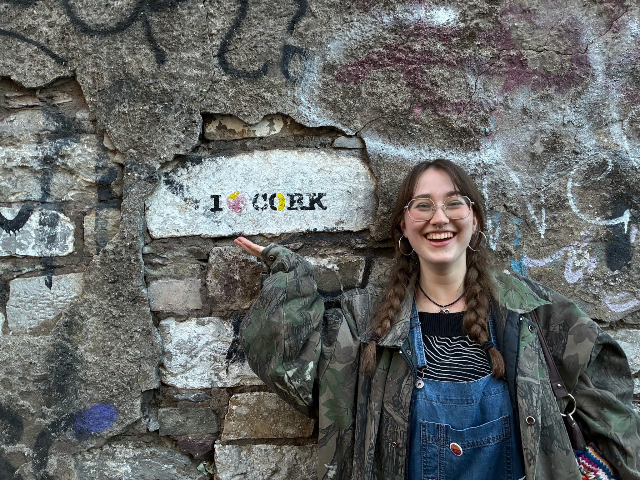 A smiling person with braided hair stands in front of a graffiti-covered stone wall, pointing at text that reads “I ♥ Cork.”.