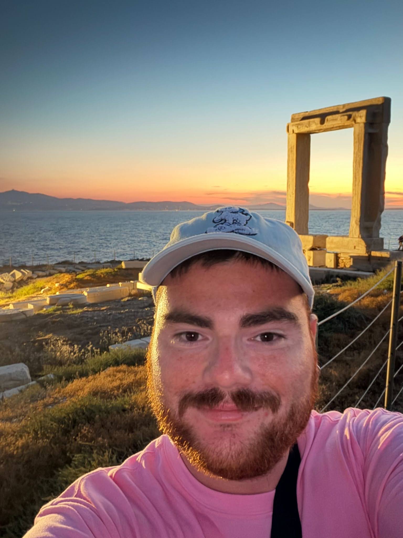 A person in a pink shirt and light cap takes a selfie at sunset near ancient stone ruins by the sea.