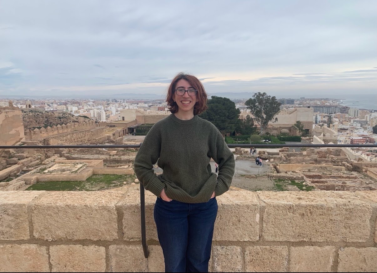 A person with glasses, wearing a green sweater and jeans, stands and smiles in front of an ancient stone wall overlooking a cityscape and distant coastline.
