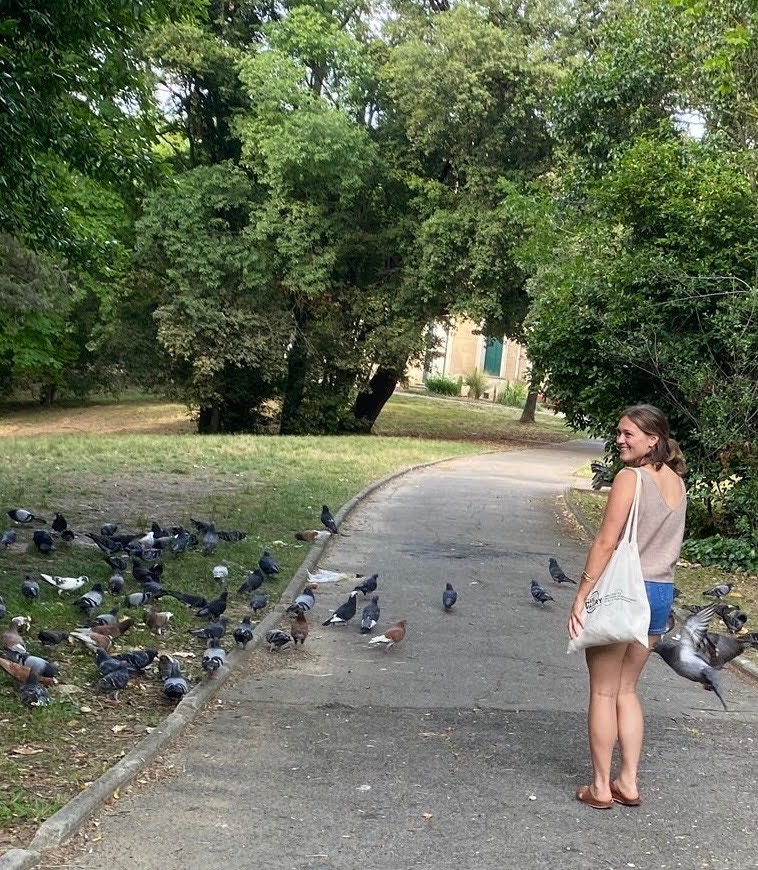 A woman with a tote bag stands on a park path, looking back at a group of pigeons gathered on the ground near green trees.