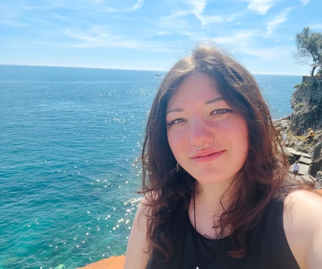 Person with long brown hair taking a selfie by a clear blue sea, with rocky cliffs and a tree visible in the background under a sunny sky.