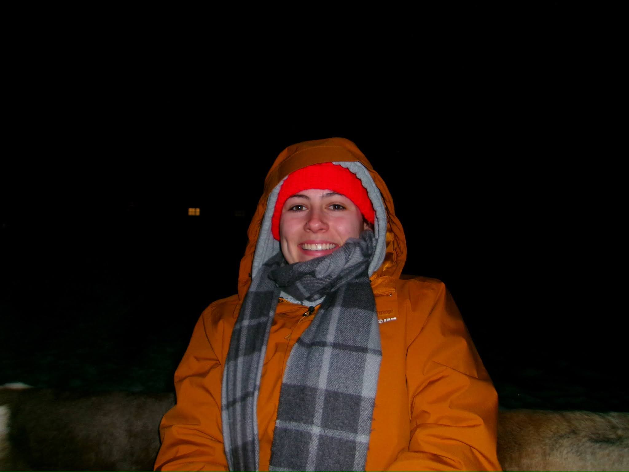 A person wearing an orange jacket, red hat, and gray plaid scarf smiles at night against a dark background.
