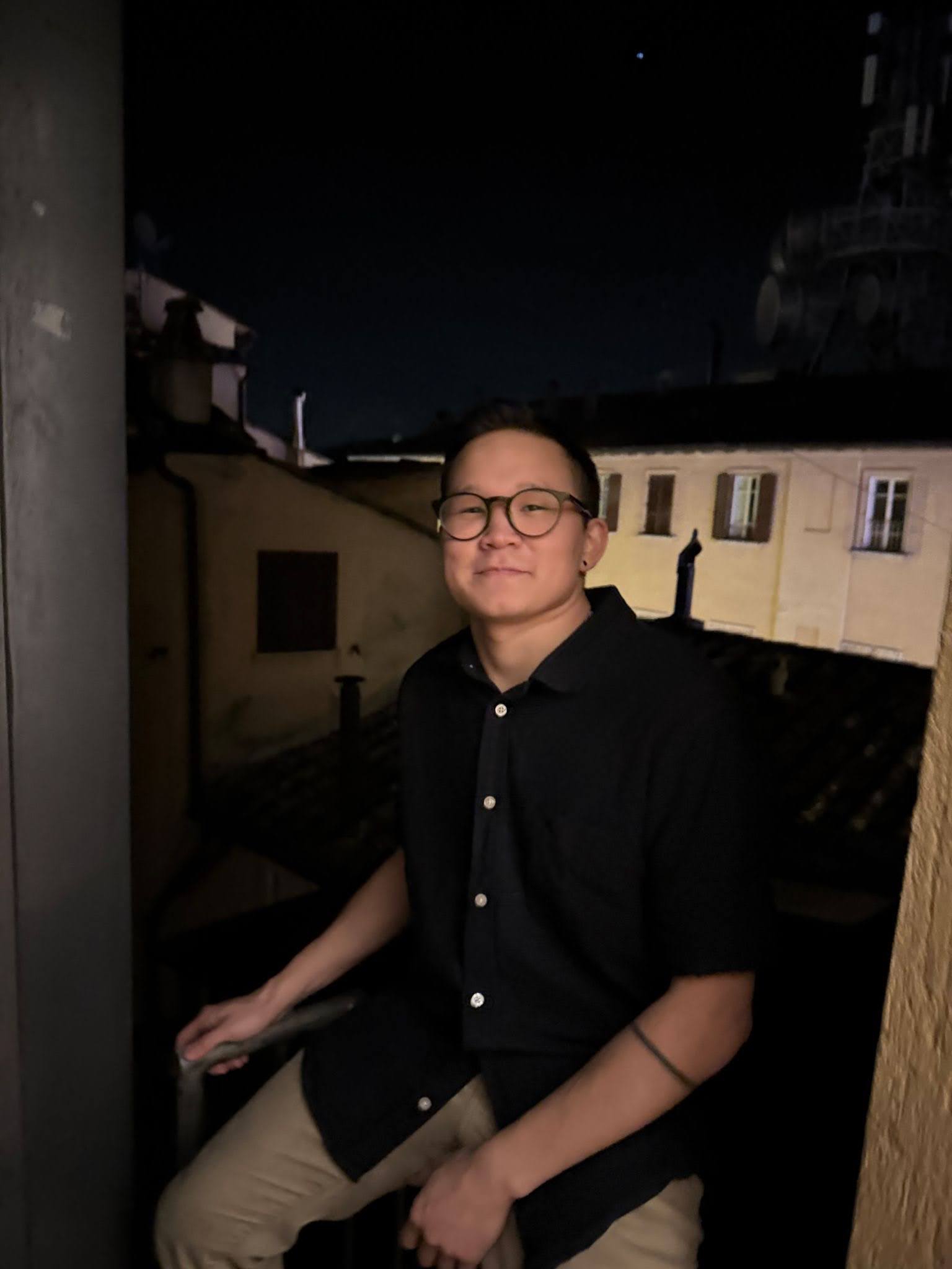A person wearing glasses and a dark shirt sits on a balcony at night, with dimly lit buildings and a star visible in the background.
