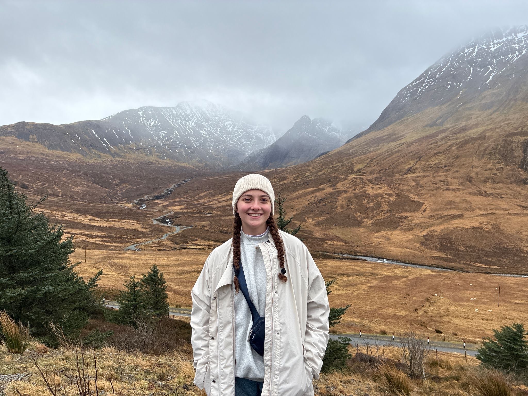 Person in a white jacket and beanie stands outdoors with brown hills, sparse trees, and snow-dusted mountains under a cloudy sky in the background.