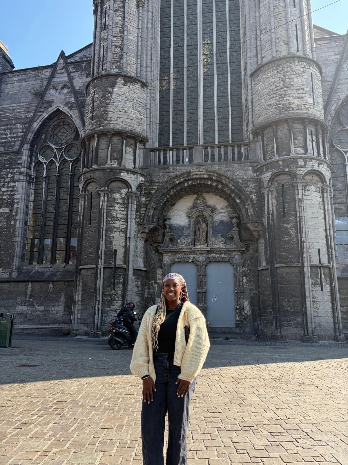 A person stands smiling in front of a large, old stone building with tall arched windows and ornate architectural details on a sunny day.