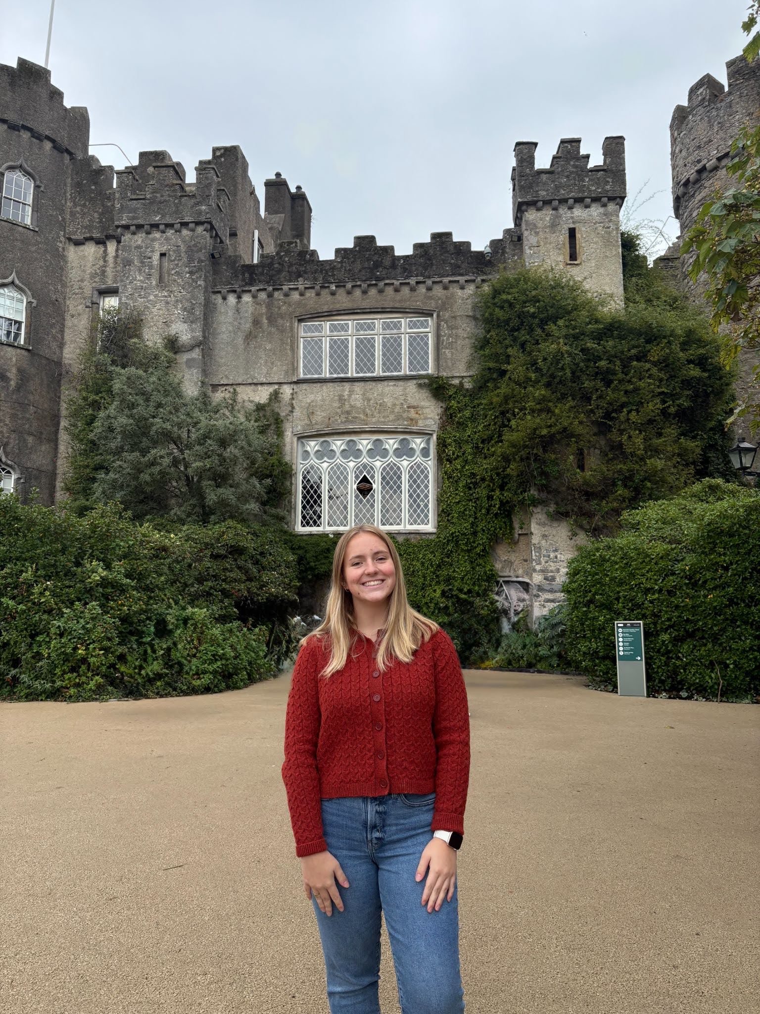 A woman in a red sweater and jeans stands smiling in front of a large stone castle with greenery and an arched window.