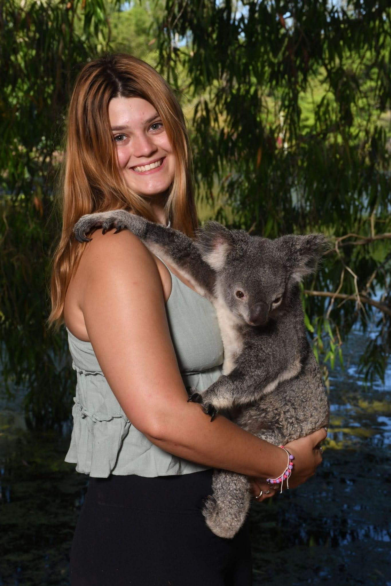 A woman with long brown hair holds a koala in her arms, standing outdoors near water and greenery.
