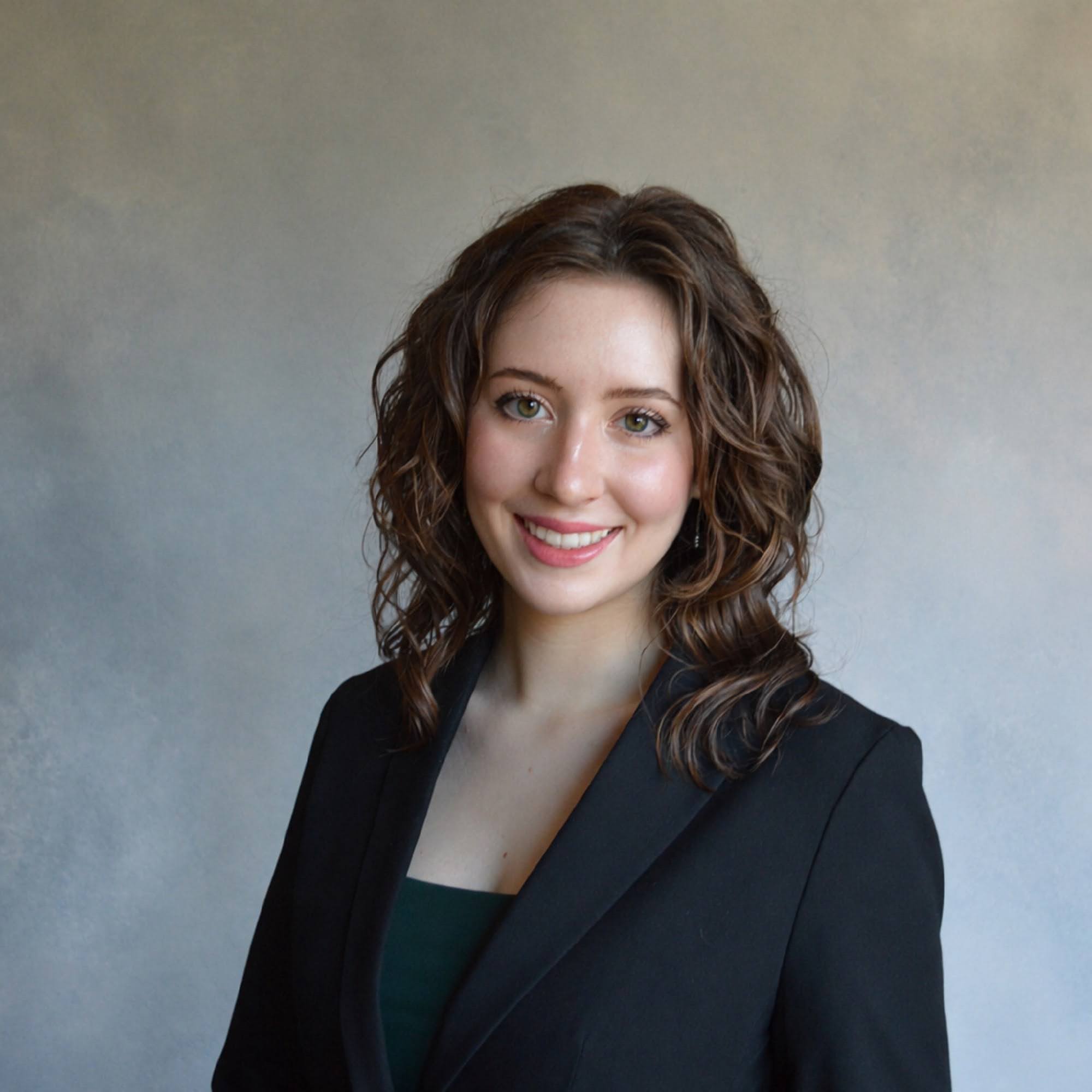 A woman with wavy brown hair, wearing a black blazer over a dark top, smiles at the camera in front of a neutral background.