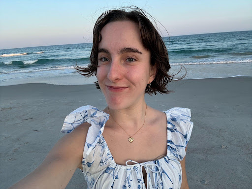 A person with short brown hair takes a selfie on a sandy beach with gentle waves and a clear sky in the background.