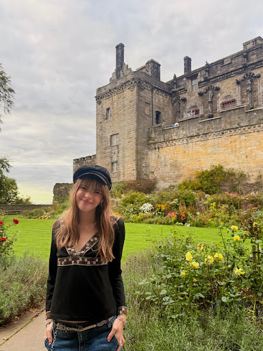 Young woman stands in front of an old stone castle with gardens and flowers, under a cloudy sky.