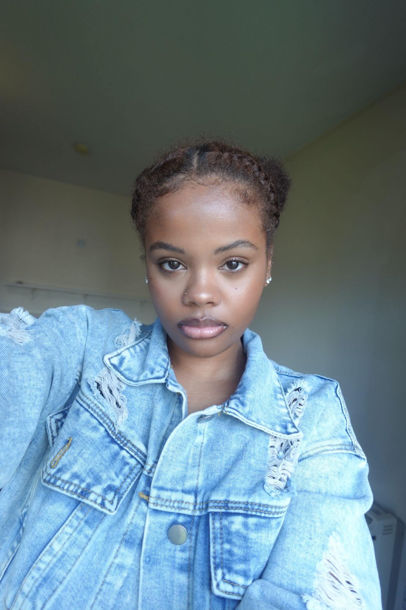 A person with braided hair wearing a distressed denim jacket looks directly at the camera with a neutral expression. The background is a plain, light-colored room.