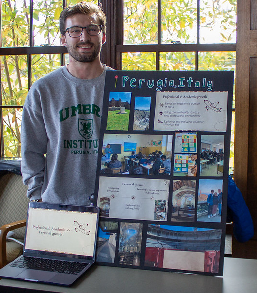 A young man standing next to a laptop and a poster board titled “Perugia, Italy” that displays photos and text about his academic and personal growth abroad.