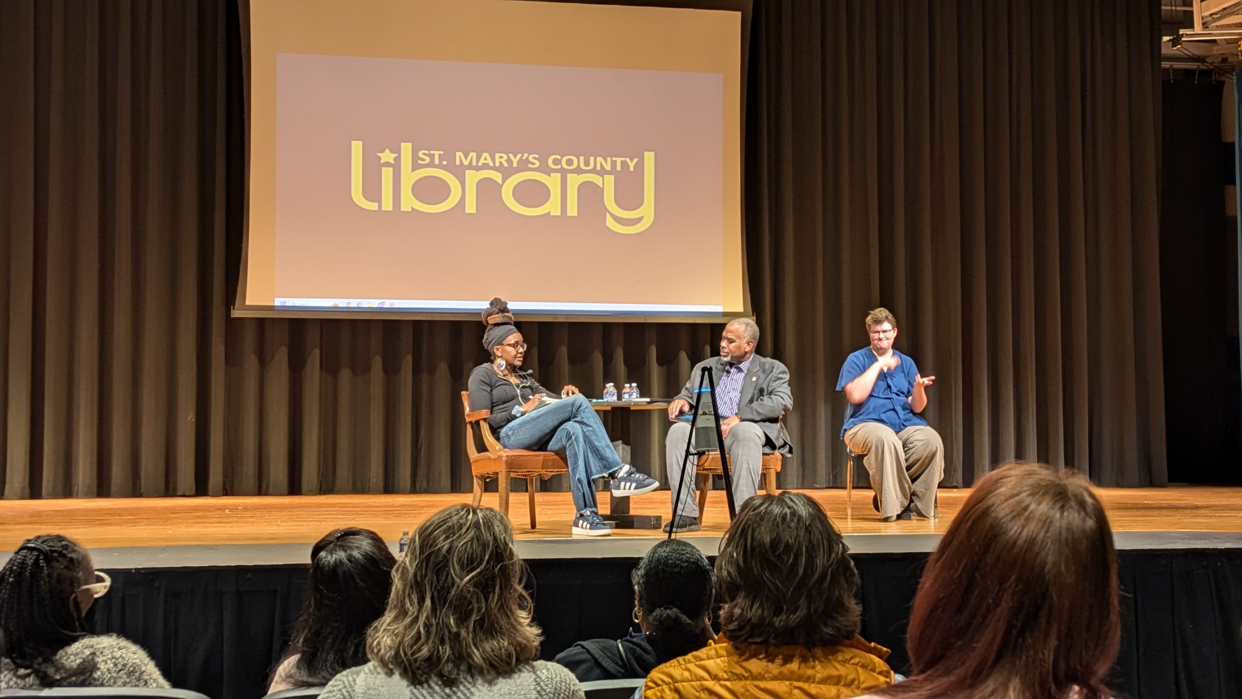 Three people sit onstage at a St. Mary's County Library event. One speaks into a microphone, another listens attentively, and the third interprets in sign language. The audience is visible in the foreground.