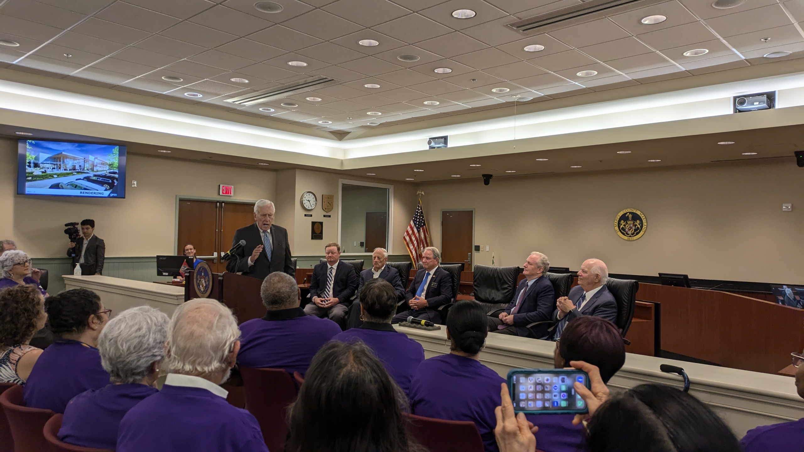 PXL_20240726_132401083 A man speaks at a podium in a courtroom, addressing an audience. Several people sit behind him, and the audience wears matching purple shirts. There's a U.S. flag and an emblem on the wall. A person in the foreground holds a smartphone.