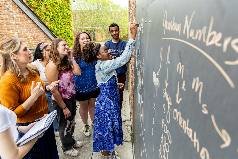A diverse group of students gather around a chalkboard outdoors. One student is writing while others watch attentively.