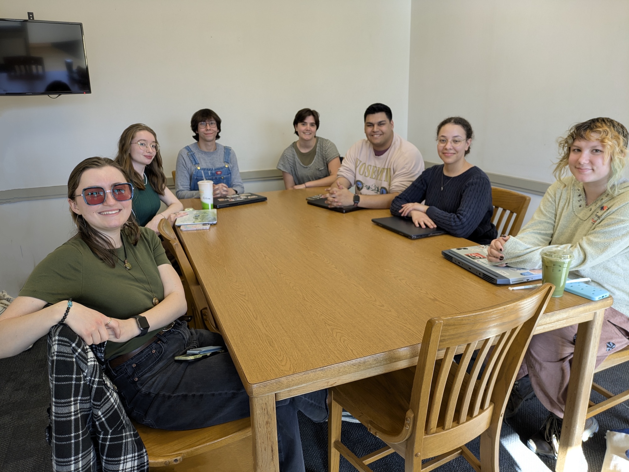 PXL_20250318_203148422~2 Seven people sitting around a wooden table in a meeting room, smiling at the camera.
