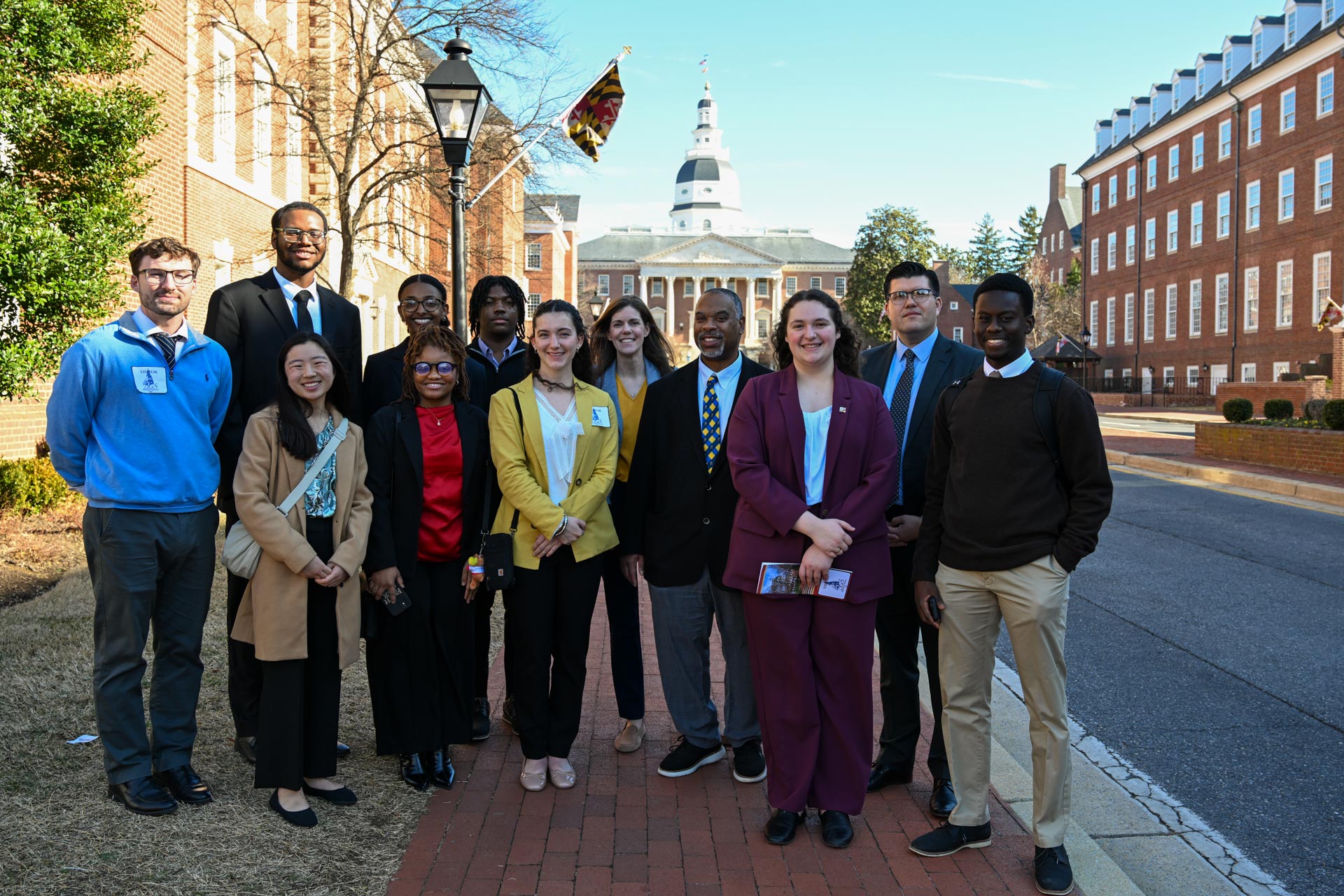 A diverse group of people stands on a brick pathway in front of a historic building with a dome and flags. They are dressed in business and casual attire, smiling at the camera, with trees and more buildings in the background.