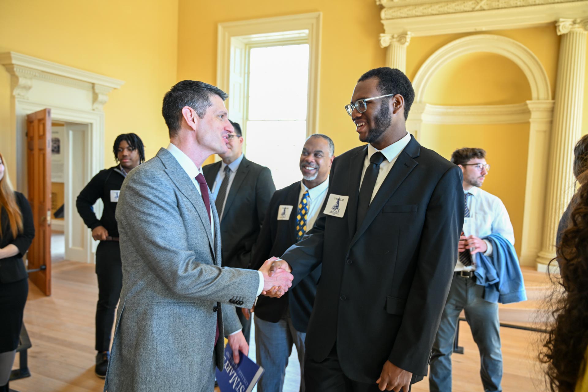 Two men in suits shake hands and smile in a room with other people and classical architecture elements. One man holds a booklet. A group of people stands in the background, engaged in conversation.