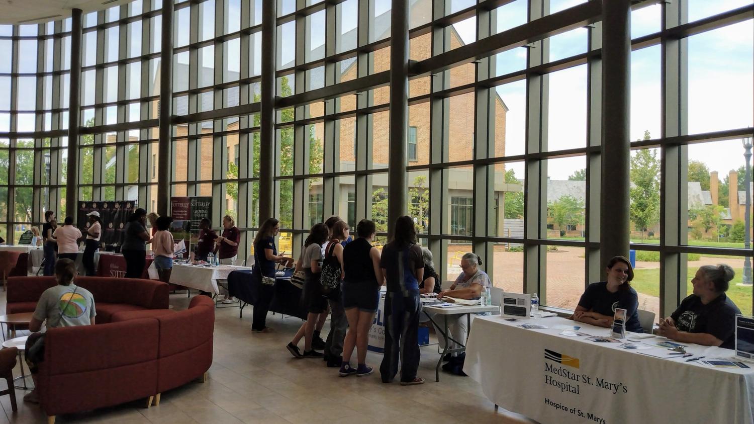 People visit information tables staffed by representatives in a spacious, window-lined lobby with views of trees and buildings outside.