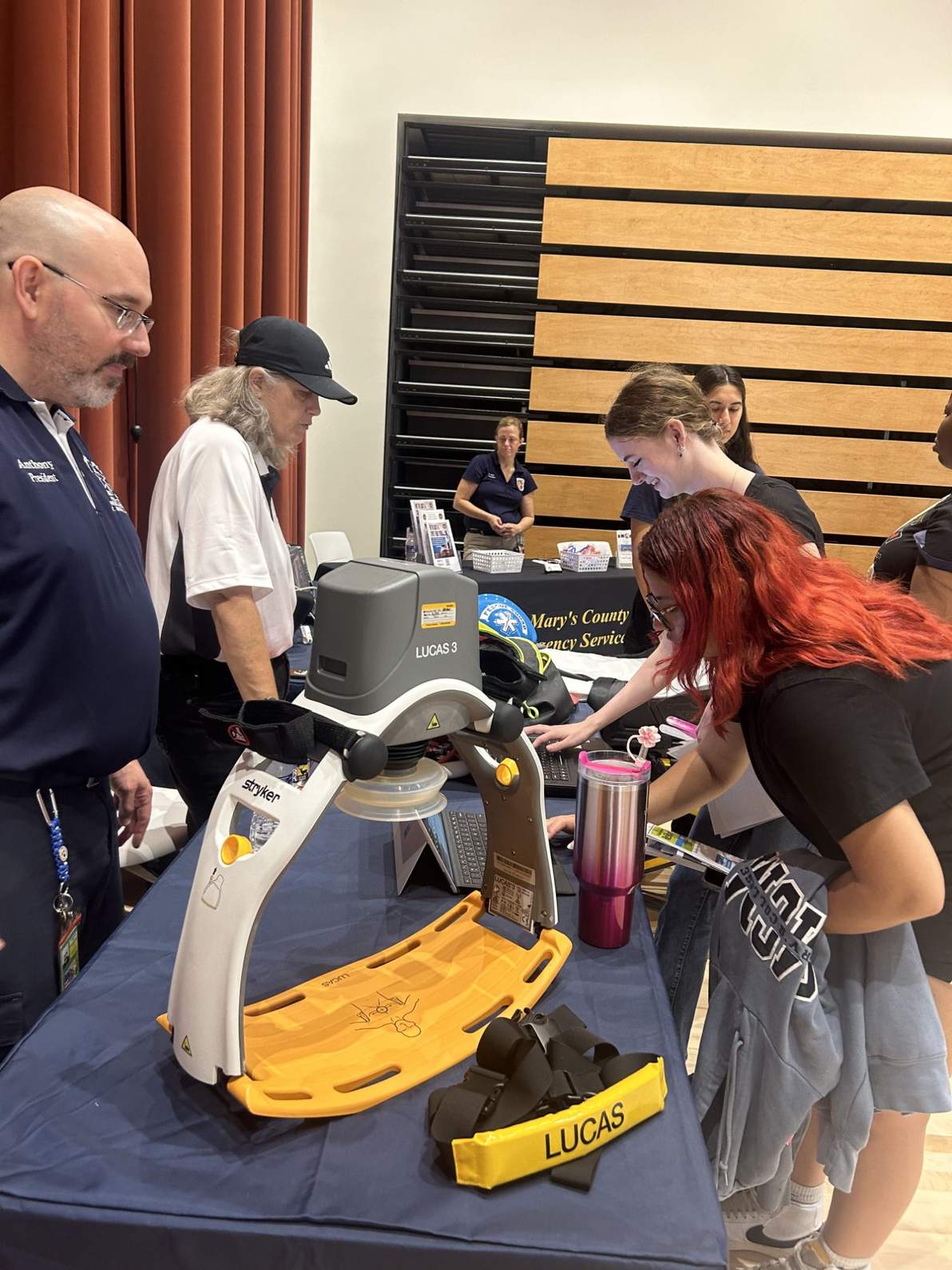 People observe and interact with a LUCAS chest compression system on display at an informational booth in a gymnasium setting.