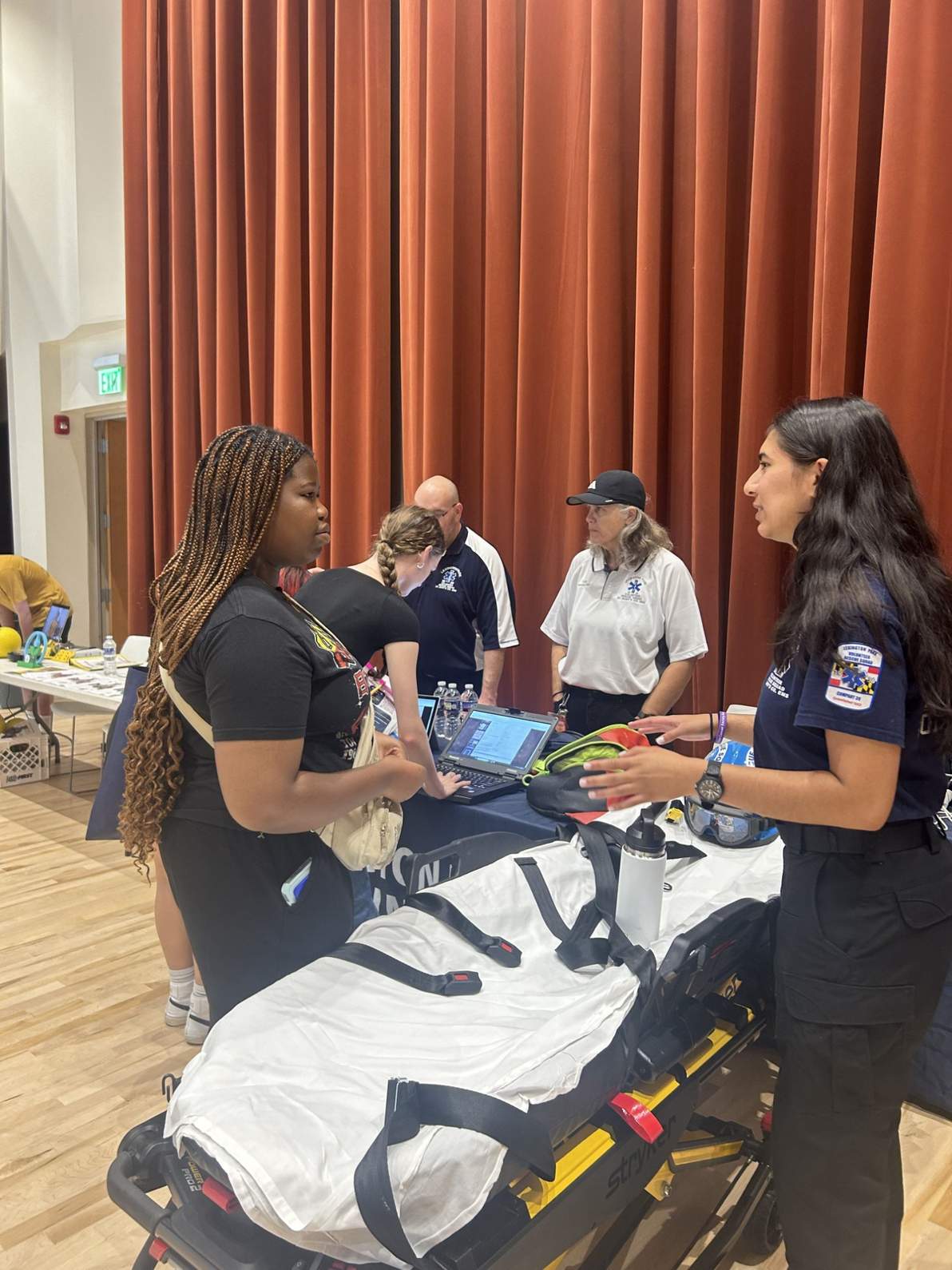 Two women converse at a medical information booth with EMS personnel; a stretcher and medical equipment are visible in the foreground.