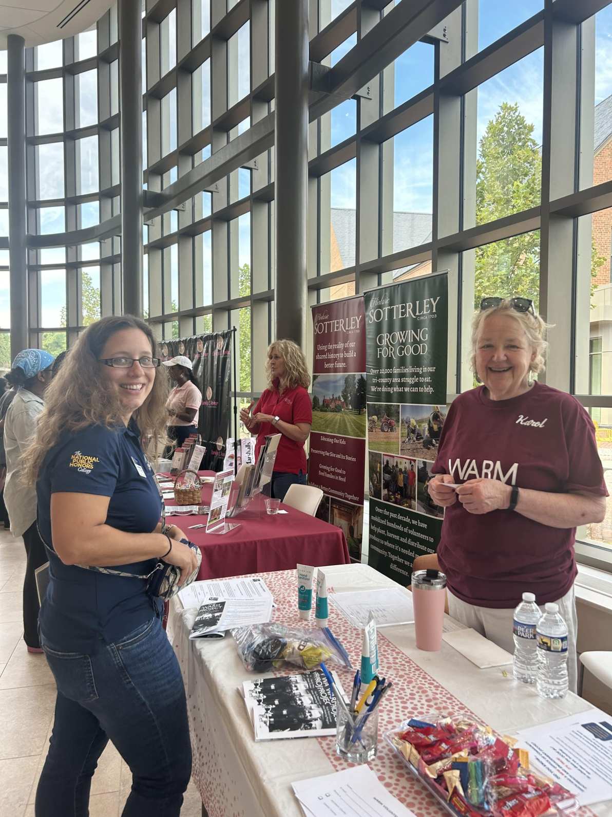 Two women stand and smile at an information table inside a brightly lit building during a community event; pamphlets and snacks are arranged on the table.