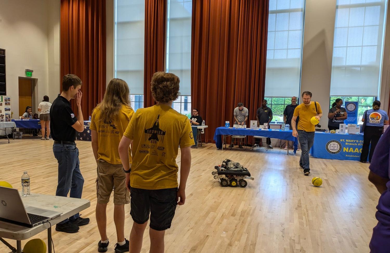 People observe and control a small robot rolling on a gym floor near a yellow ball, with informational booths and attendees in the background.