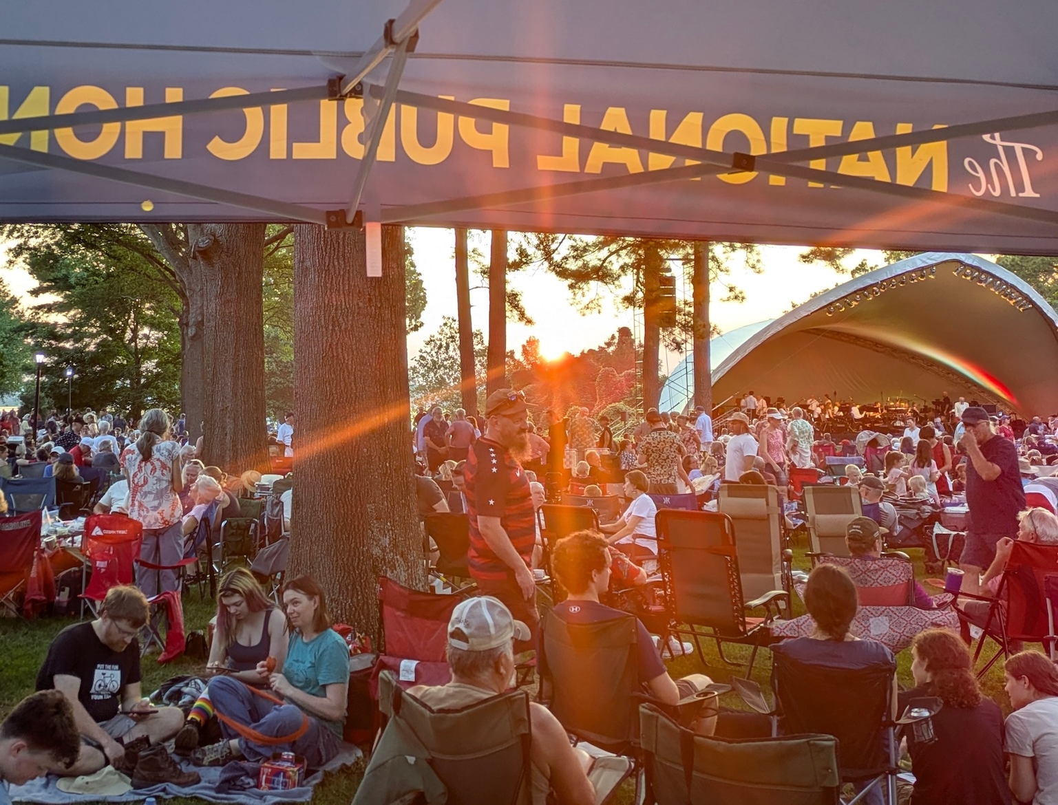 Crowd seated on lawn chairs and blankets at an outdoor concert near an amphitheater, viewed from under a tent with text partially visible on the canopy.