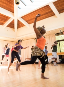 A group of people participate in an energetic dance class in a sunlit studio with wooden floors and a high ceiling.