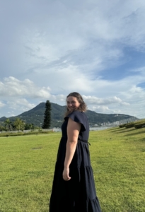 A person in a black dress stands on a grassy field, smiling at the camera.