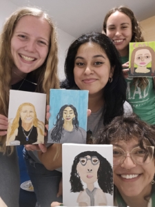 Four smiling people hold up their own painted self-portraits. Each portrait shows different styles and colors.