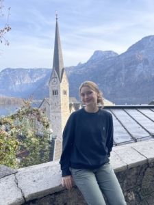 A person in a black sweater leans against a stone wall with a picturesque background of a church steeple, mountains, and a lake under a blue sky.