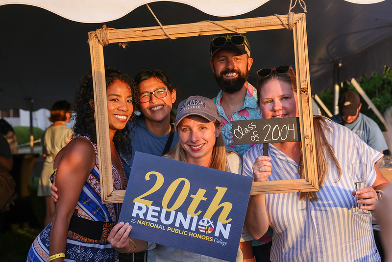 Five people pose together at an outdoor event, holding a large wooden frame and signs that read "Class of 2004" and "20th Reunion National Public Honors College.