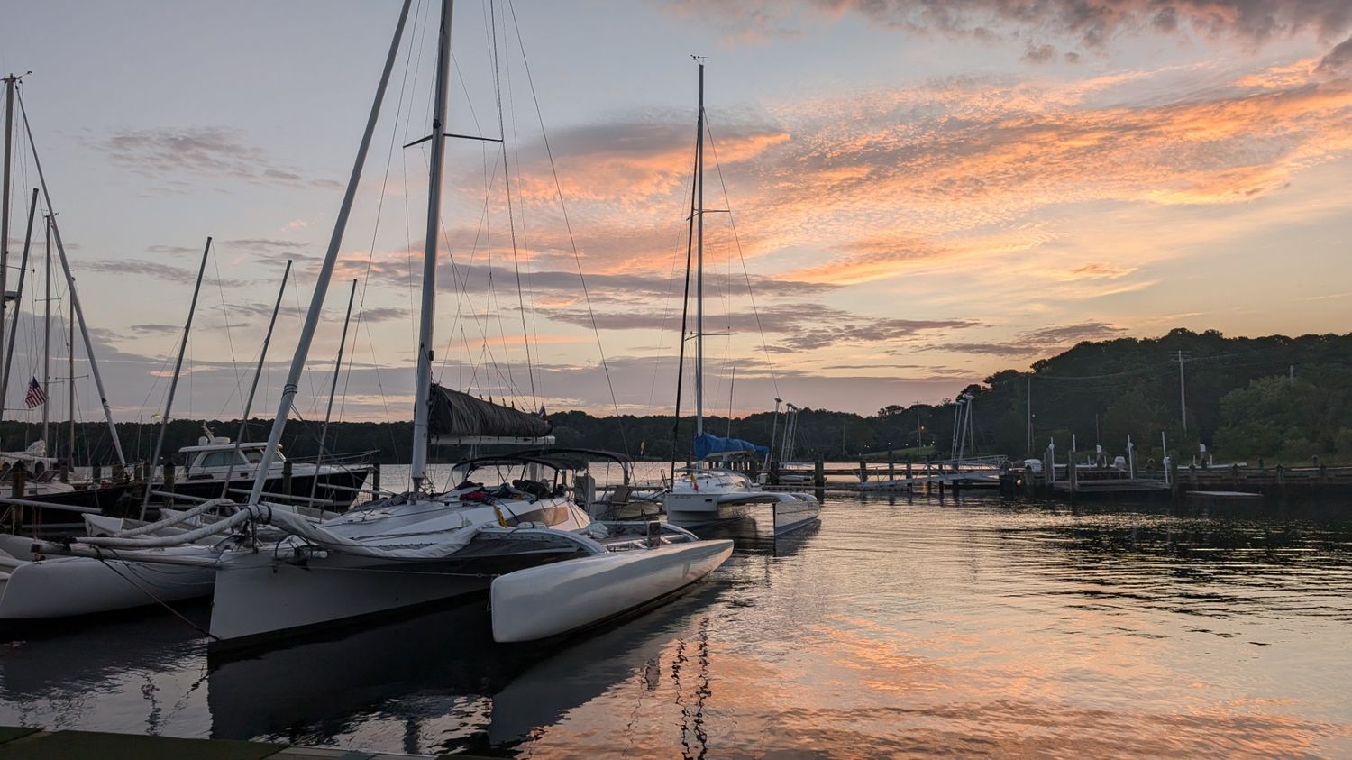 Sailboats docked at a marina during sunset, with calm water reflecting the sky and wooded hills in the background.