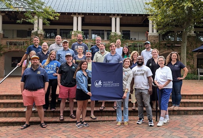 A group of people stand outdoors on brick steps, smiling at the camera and holding a banner that reads "Alumni Association." A building with a balcony is visible in the background.