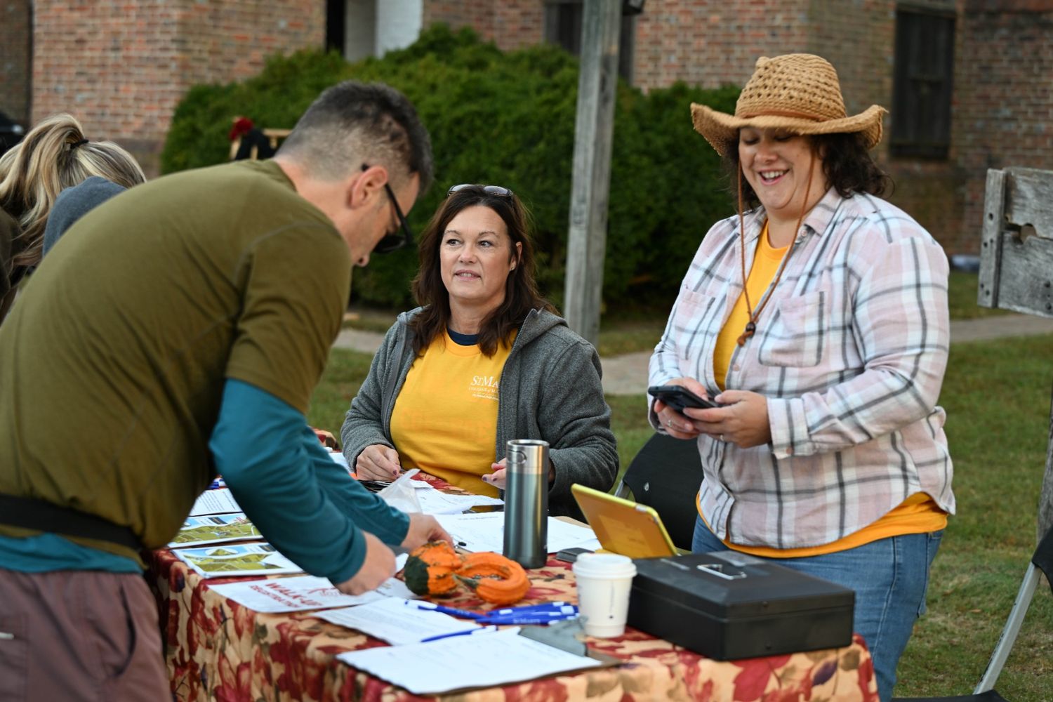 Two women sit at a table with papers and a cash box outdoors, talking to a man filling out a form. One woman is smiling and wearing a straw hat.