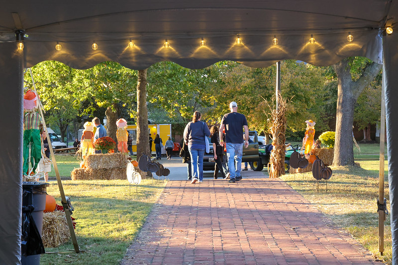 People walk along a brick path at an outdoor event decorated with scarecrows, hay bales, and string lights in the evening.