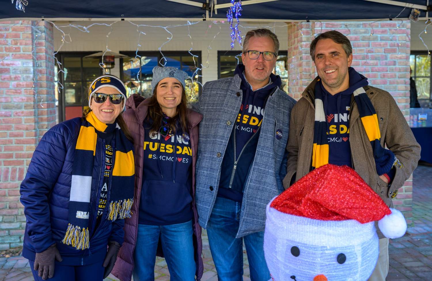 Four adults in winter clothing and "Giving Tuesday" shirts pose together outdoors beside a snowman decoration under a canopy with string lights.