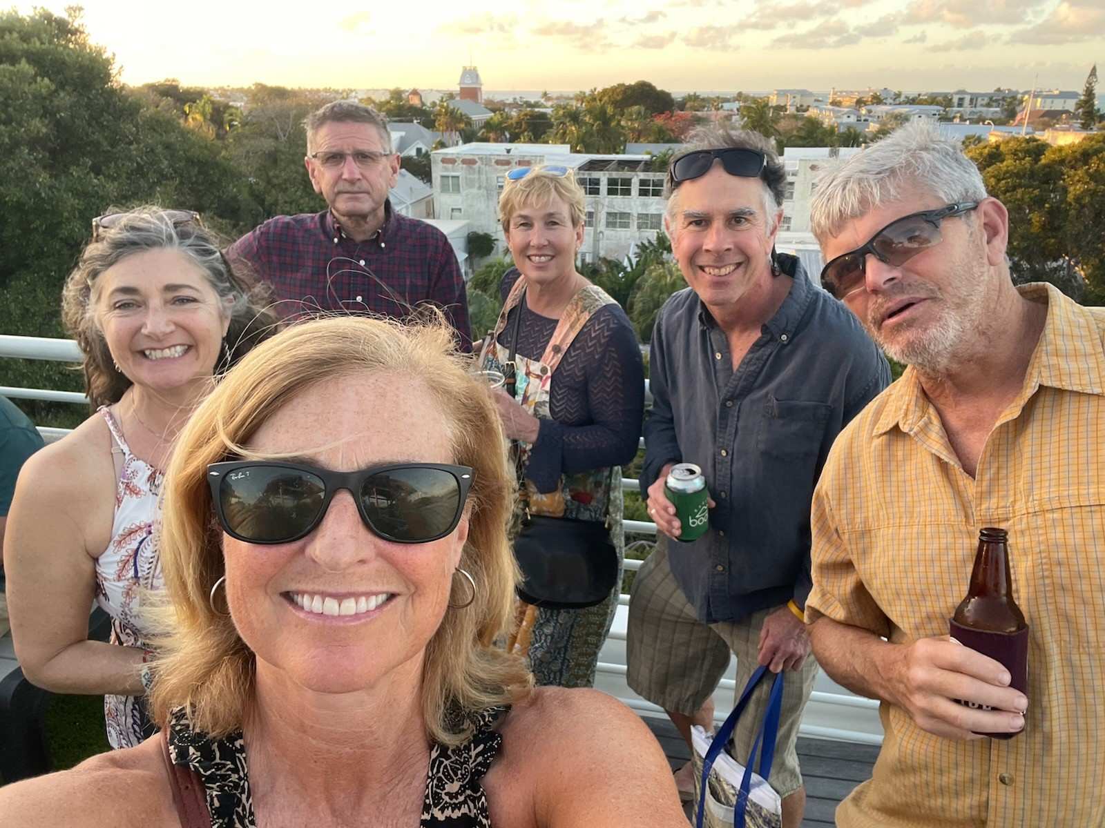 Six adults stand together outdoors at sunset, smiling at the camera; two hold beverages, and trees and buildings are visible in the background.