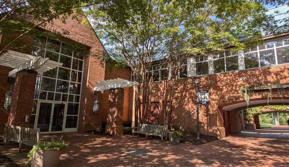 Red brick building with large windows, leafy trees casting shadows, benches along the wall, and a covered archway leading to a walkway in the background.